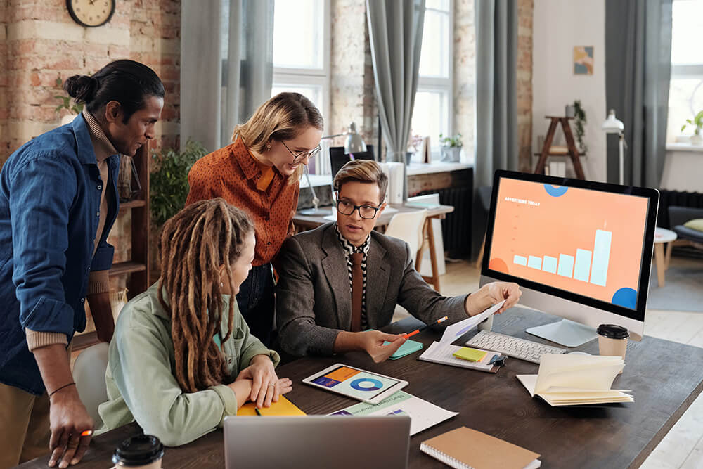 A Group of People Discussing Beside a Desktop with Graph on Screen.